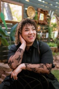 Noelle Monét, a Black-biracial woman with curly brown and blond hair, sits outdoors, backwards on an iron chair, she's wearing a black collared top with black lace sleeves, and rests her head in her hand while looking off to the side with a wide smile.