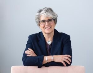 Author photo of Oona Metz wearing a blue jacket, with arms folded leaning on a pink chair.