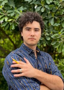 P.C. Verrone, a man with curly brown hair wearing a blue collared shirt, stands in front of a green bush. He is holding a pencil and looking at the viewer.