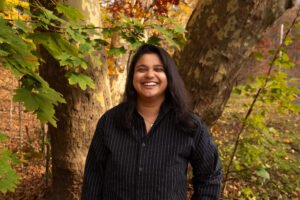 Padya Paramita, a Bangladeshi person with shoulder length hair, stands in the park in a black button down shirt.