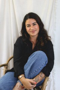 RJ Valldeperas, a Latina woman with long dark hair, sits in a chair with one leg up and her arms bracing her leg. She is looking at the camera with a small smile.