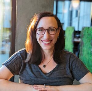 A white woman with brown hair and burgundy glasses smiles.