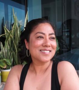 Photo shows a smiling brown woman with shoulder length black hair and a black tank top. She is show from the chest up against a background of glass and plants.