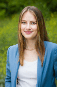 Rebecca Bendheim, a white woman with blonde/brown hair, is wearing a light blue blazer with a background of greenery.
