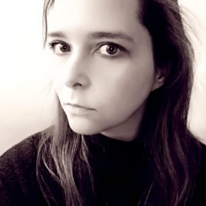 Rebecca A. Carter, a white woman with long brunette hair in a half up half down hairdo. Picture is black and white, close up distance, and the author is looking sideways to the camera with a small smile.