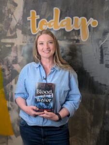 A blonde woman in a blue shirt and jeans is smiling and holding a copy of her book - Blood and water.