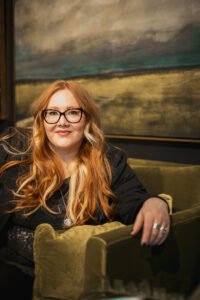 Rebecca Sky, a white person with long red hair and glasses wearing a green lace shirt and black jacket and sitting on a green velvet couch. Rebecca is looking at the viewer with a cheeky smile.