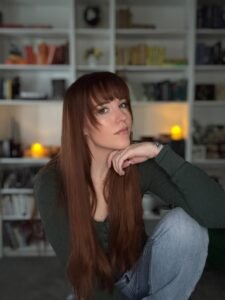Revey Galaer, a white woman with fringe bangs and long red hair, sits on a stool before a wall of white bookshelves. She rests her chin on her hand as she stares into the camera.
