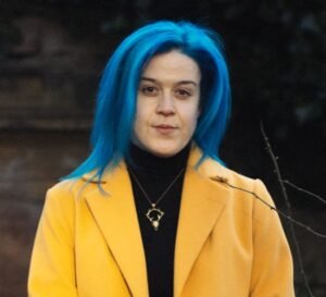 Rhiannon Grist, a white cis woman with shoulder-length blue hair, is outdoors in an autumnal graveyard. She wears a black polo neck and a yellow coat. She is looking at the viewer with a serious expression.
