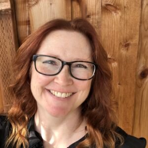 A photo of Rie Neal, a white woman with wavy red hair and glasses, wearing a black blouse with short sleeves. A wooden fence is in the background. Rie is smiling at the camera; only her head and shoulders are shown in the picture.