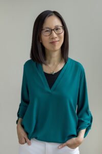 Roberta Kwok, an Asian woman with shoulder-length black hair wearing a blue dress shirt, white pants, and glasses. She has her hands in her pockets and is smiling.