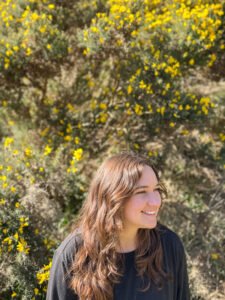 Robyn J Pritzker, a white woman with wavy brown hair, sits outside in the sun in front of some marigold gorse. She is looking off to her left, smiling.
