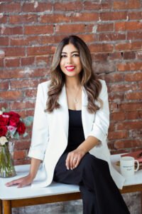 Rosey Kaur Hwang, a South Asian woman with long dark highlighted hair, is sitting on a desk with roses next to her, smiling.