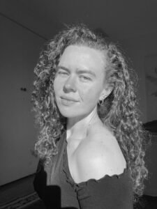 Rosie Sycamore, a white woman with curly hair and wearing a black t-shirt. She is sitting side-on, looking at the camera with a slight smile.