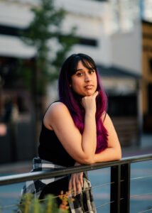 A photo of Indian-American author S.C. Bandreddi. She is looking at the camera as she leans against a railing with her chin on her hand. Her ombré black and purple hair reaches her other arm which is resting on the railing. She is wearing a black tank top and has a gray and white flannel wrapped around her waist.