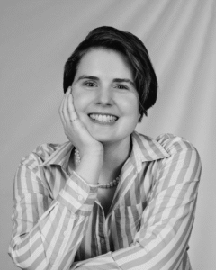 A black and white photograph of a white woman with short dark hair, holding her chin in her hand. She is wearing a ring on her pink, a double strand of pearls, and a striped button down blouse. She is smiling widely.