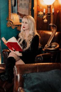 Sarah Glenn Marsh, a white woman with long, wavy blond hair and red lipstick, holds a book and stares off into the distance while sitting beside a large gold cobra statue.