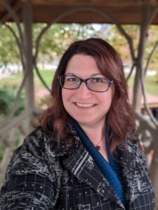 White woman with dark reddish brown hair is wearing a black coat with map patterning, standing smiling in front of wooden circle designs of a gazebo. She wears glasses and is looking straight at the camera.