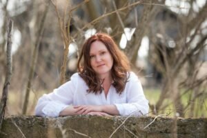 Sarah Van Goethem, a white woman with tousled auburn hair, stands outdoors in nature with her arms on an old stone crumbling fence. She has a slight smile on her face.
