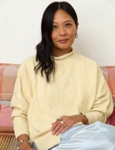 Sarah Wang, a Chinese woman with long black hair, sits on a sofa with one arm across her lap. She is smiling and looking directly at the viewer.