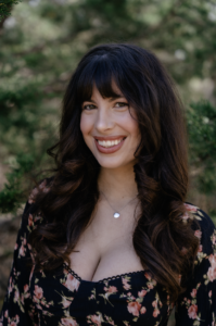 A brunette woman wearing a black and pink floral dress is smiling at the camera. She is standing outdoors among dark green trees.