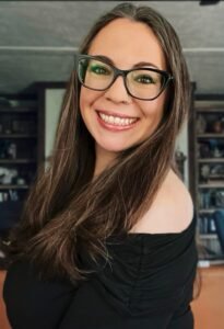 Sheena Holbrooks/pen name Arien Skye, a white woman with long brown hair and black rimmed glasses smiles wide in front of blurred bookshelves.
