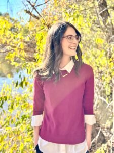 Sierra Branham, a white woman with medium length brown hair, stands outdoors, staring off to the side, smiling and backlit by the sun.
