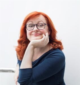 A redheaded middle-aged white woman looks quizzically at the camera, her chin resting on her hand.