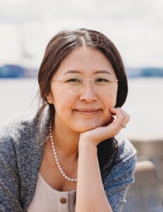Sophie Li, an Asian woman with long dark hair, sits outdoors with her hand cradling her chin. She is looking at the viewer and has a bright smile.
