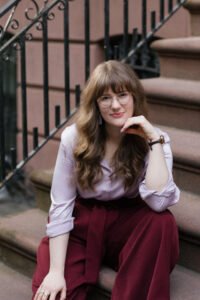 Sophia Huneycutt, a white woman with long, wavy light brown hair, sits on the front steps of a brownstone, hand under her chin. She is looking at the viewer and smiling at the camera. She wears gold, round glasses, a lavender silk button-down and burgundy wide-leg trousers.