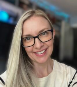 A close-up head-shot of Sophie L. Henderson, a white woman with long straight blonde hair and black rimmed glasses. She is inside, with a blurred background, looking at the viewer, and is smiling broadly.