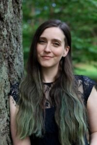 A white woman with long brown and green hair wearing a black lacy dress stands against a tree in a forest.