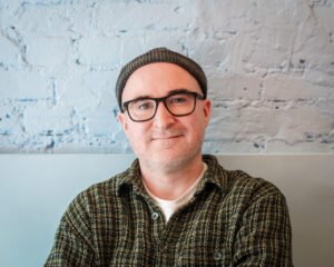 A man with glasses in a blue dockers cap and green shirt stands in front of a light blue painted wall with exposed brick.