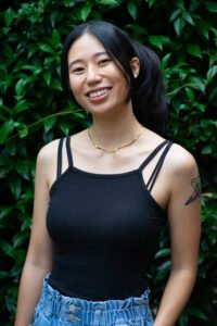 A Chinese woman with her hair pulled into a pony tail wearing a black tank top standing in front of lush greenery. She is smiling at the camera.