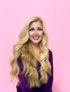 TC Kemper, a white woman with very long, blonde hair, sits in front of a pink background. She is smiling.