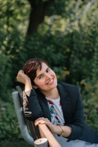 A smiling woman with short black hair and olive skin, seated on a park bench.