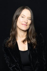 Tara Goedjen, a woman wearing a black blazer with dark brown hair, just past her shoulders, looking at the viewer with a smile.