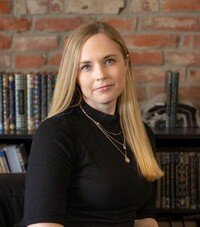 A white female with blonde hair in a black sweater with a rose gold necklace sits in front of a brick wall and bookshelf.