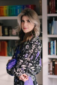 An image of Taylor Grothe in a purple mushroom patterned top against a background of rainbow books. They are looking at the camera 3/4 on and smiling slightly.