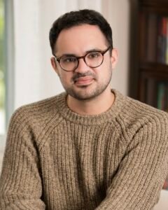 A white, 30s male author with short, dark brown hair, round tortoise-shell glasses, and a light-brown sweater sits in front of a window and a bookcase.