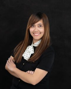 Photo of Trish Caragan, a Filipina woman with long brown hair and side swept bangs. She is wearing a black dress with gold buttons and a white and black polka-dotted collar tied in a bow. She's standing in front of a dark background and wearing pinkish purple lipstick. Her arms are crossed and her nails are royal blue. She is, like her book, SMILING for the CAMERA!