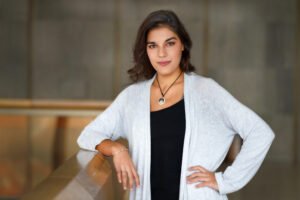 Via Luino, a woman with Mediterranean skin tones, dark brown hair, and dark brown eyes, smiles subtly at the the camera. Her right arm is propped on a wooden railing, her left hand on her hip. She wears a white cardigan over a black top and a necklace of a wolf howling at a shooting star.