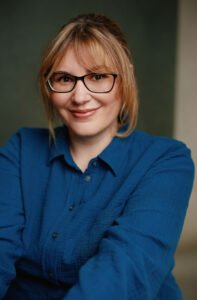 V.A. Vazquez, a white woman with blonde hair styled in a low updo, wearing black glasses and a textured blue blouse, smiling warmly at the camera against a softly blurred gray backdrop.