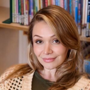 Violet Lumani, a white woman with dark blonde hair, sits indoors with a hunter green shirt and beige crochet sweater. She is looking at the viewer and has a slight closed mouth smile on her face.