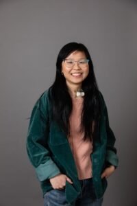 Wendy Lu, a Chinese American woman with long black hair, is standing with her hands partly in her pockets. She is wearing glasses, a tracheostomy tube, and a corduroy button-down jacket over a pink sweater.