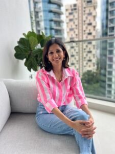 Yashika Mody, an Indian woman with medium black hair, is wearing a neon pink and white striped shirt and blue jeans. She is sitting outdoors in a balcony on a grey sofa in front of a green plant. She is looking at the viewer with a joyful smile.