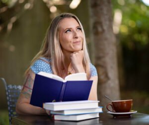 A blonde woman with a blue polka-dotted shirtholds a book and looks off into the distance, smiling.